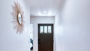 Käytävä, jossa on puulattia, valkoiset seinät, tumma ovi ja auringonsäteitä muistuttava peili vasemmalla seinällä.