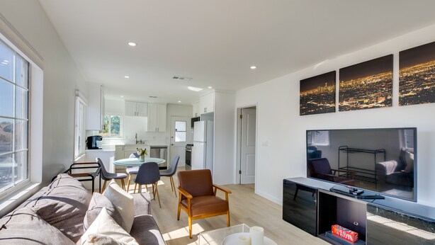 En moderne stue med sofa, sofabord og fjernsyn. I baggrunden er der et køkken med hvide skabe og et spiseområde med stole.