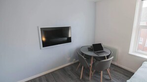 Ein kleines Zimmer mit einem flachbildfernseher an der Wand, einem runden Tisch mit einem Laptop und einem Bett mit heller Decke.