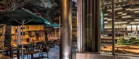 An outdoor seating area with tables and chairs, a large pillar, and a view into a modern interior space with a bar and seating.