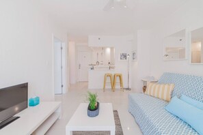 Una sala moderna con un sofá, una mesa de centro y un televisor. Se observa un área de cocina al fondo, con taburetes y una barra.