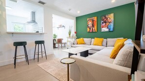 Una sala de estar moderna con un sofá, dos taburetes de bar, una mesa de centro y una pared verde decorativa con obras de arte.