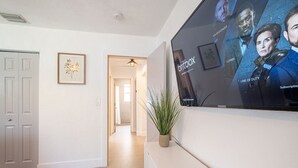 Una sala moderna con un televisor de pantalla plana montado en la pared, una planta en una mesita de consola y un pasillo que conduce a otra habitación.