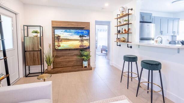Una sala moderna con un televisor grande, una pared decorativa de madera, una zona de cocina con taburetes de bar y vistas al exterior.