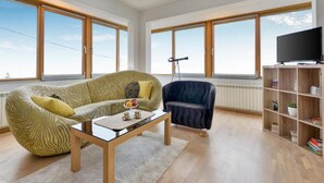 A spacious living room with a zebra-patterned sofa, a dark armchair, a glass coffee table, and a bookshelf. Large windows provide ample natural light and views.