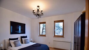 A bedroom with a bed, a chandelier, two windows, a radiator, and a wardrobe.
