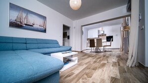 A modern living room with a blue sofa, a white coffee table, and a painting of sailboats on the wall. There is a kitchen visible in the background with a dining area. - Villa Gulli - Apartment Blue with Sea View (Supetarska Draga)