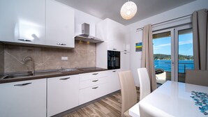 A modern kitchen with white cabinets, a stainless steel sink, and a built-in oven. There is a dining area with a white table and chairs, and a view of the sea through a large window. - Villa Gulli - Apartment Blue with Sea View (Supetarska Draga)