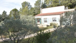Une maison à deux étages, avec un toit recouvert de tuiles rouges, entourée d’oliviers et bordée d’un chemin de gravier.
