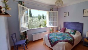 Une chambre avec une grande fenêtre, un lit doté d’une tête de lit violette et un fauteuil.