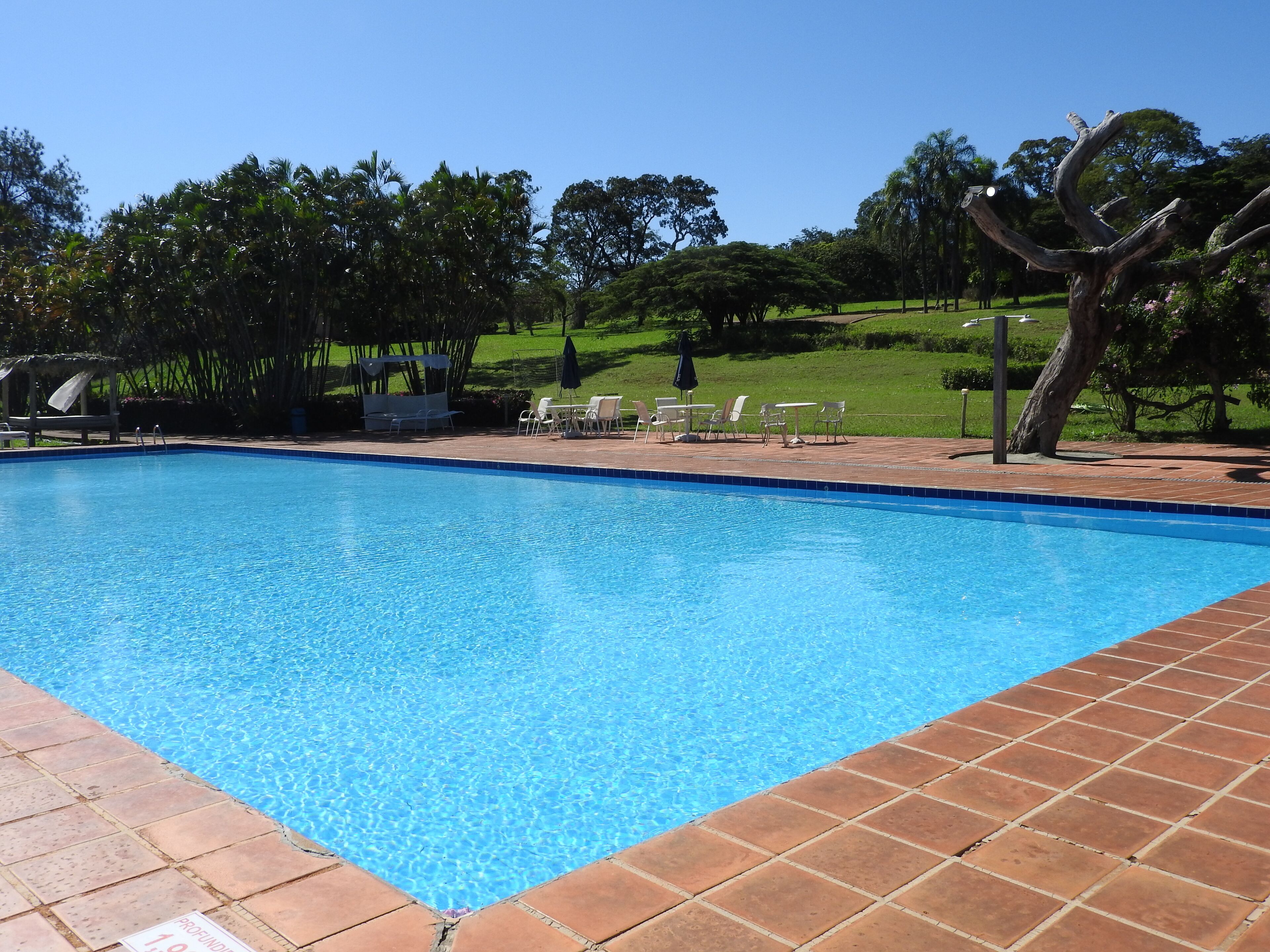 Uma piscina com bordas azulejadas, rodeada por um alpendre com cadeiras e uma pérgula.