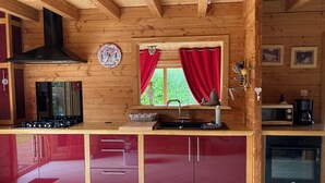 Une cuisine rustique, située dans une cabane en bois, équipée d’une cuisinière à gaz, d’un four à micro-ondes, d’un évier et d’un rideau rouge devant une fenêtre.