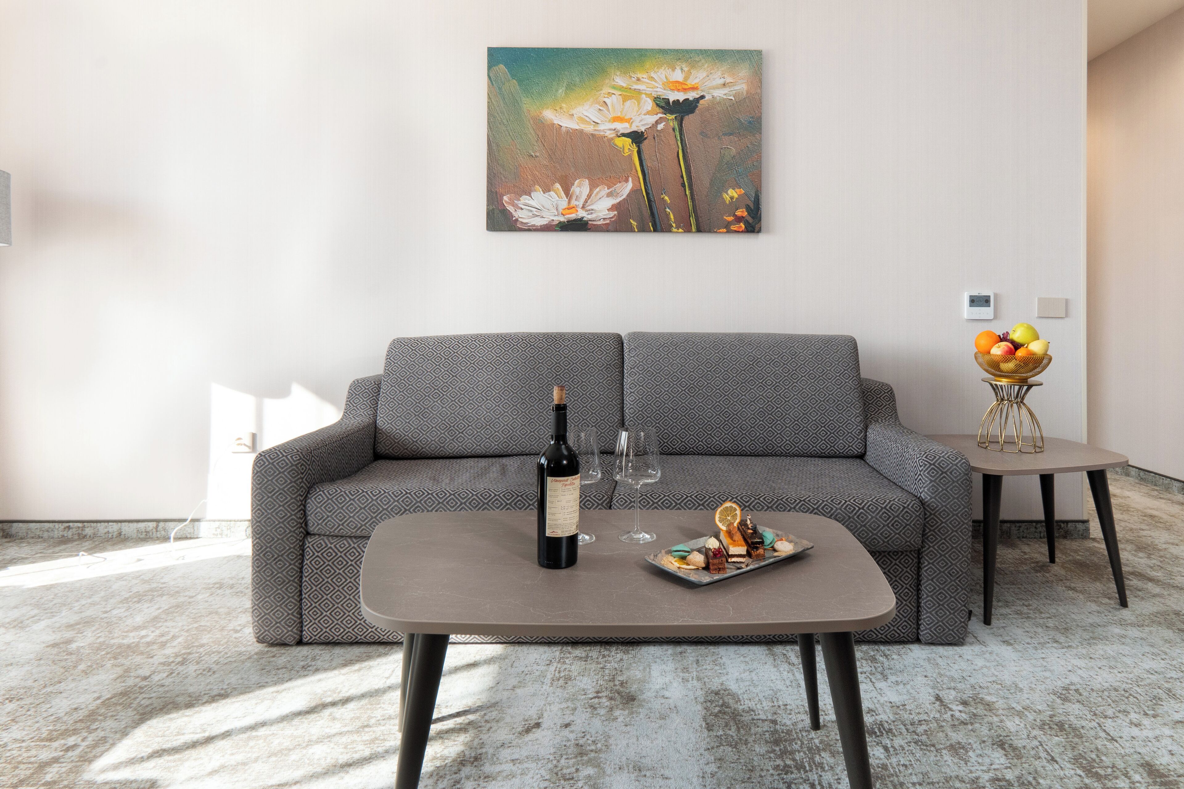 A modern living room with a grey patterned sofa, a coffee table with a bottle of wine and glasses, and a painting of flowers on the wall.