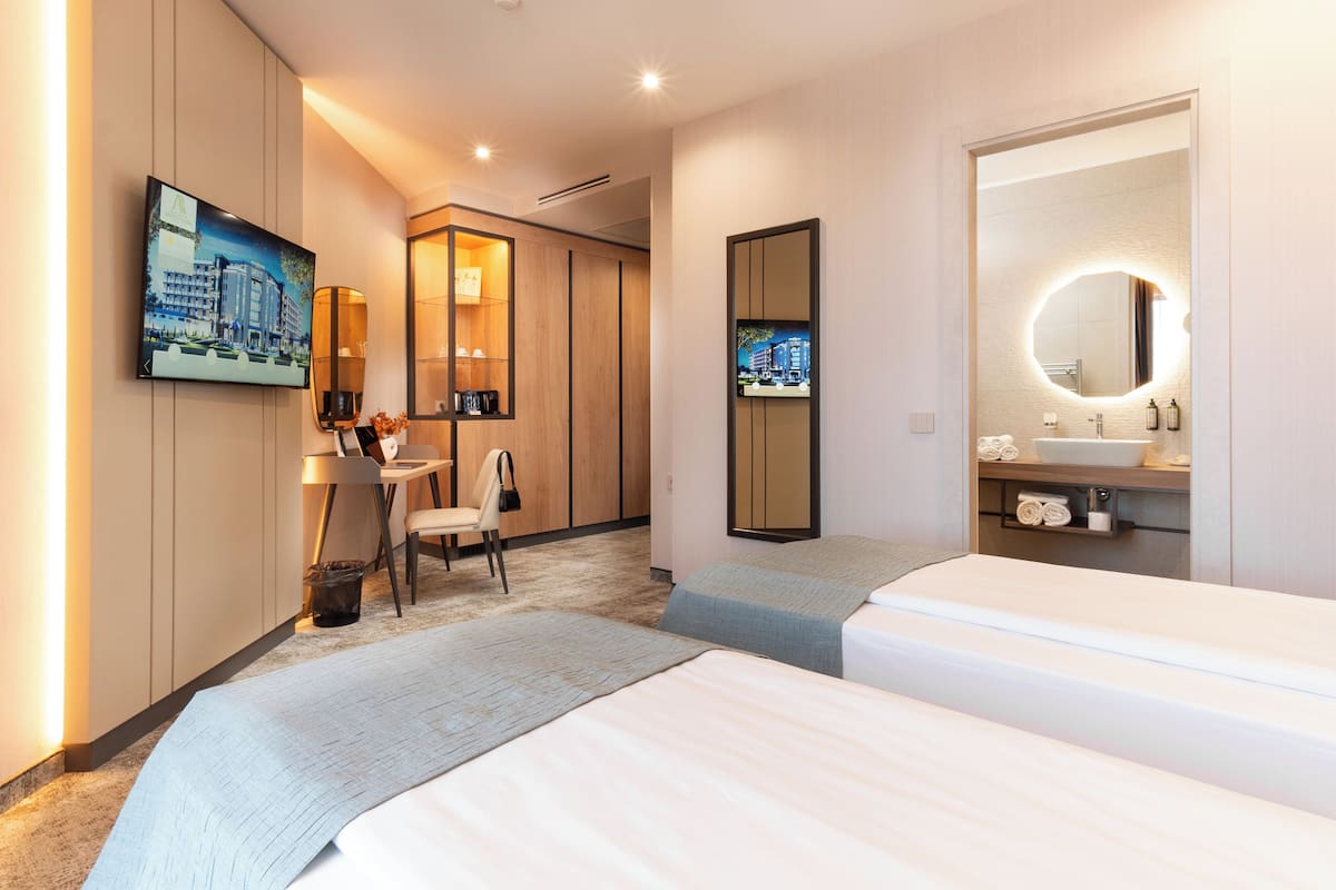 A hotel room with two beds, a desk with a chair, a large mirror, a TV mounted on the wall, and a bathroom with a sink and mirror.