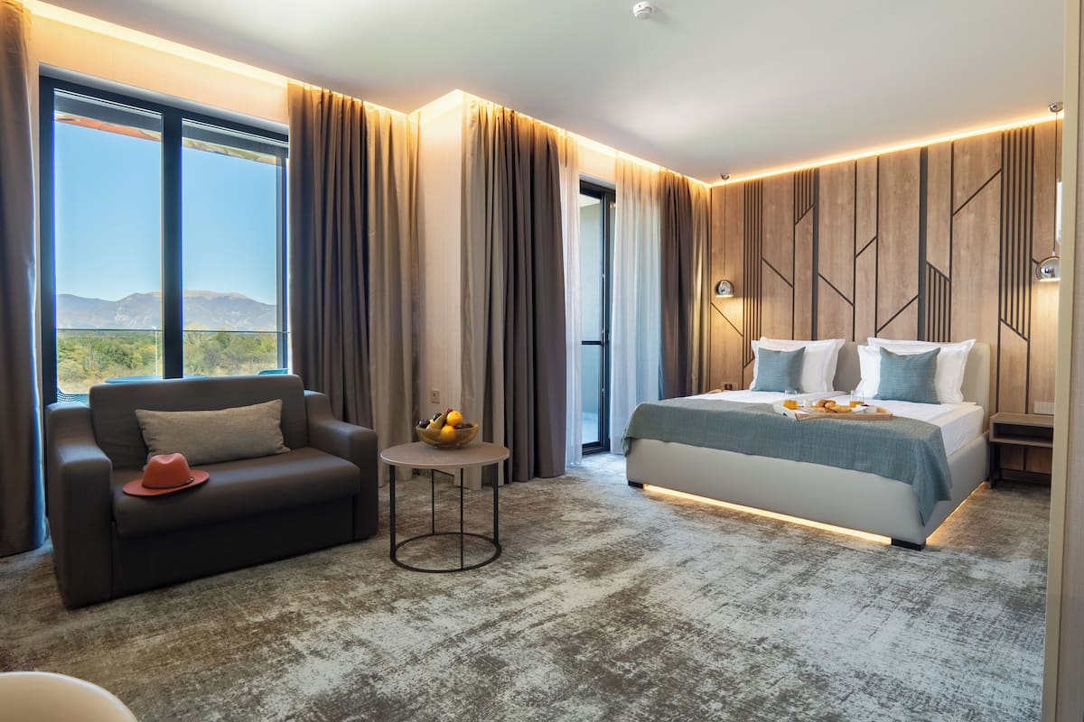 A modern hotel room with a large bed, a sofa, a small table with fruit, and a view of mountains.