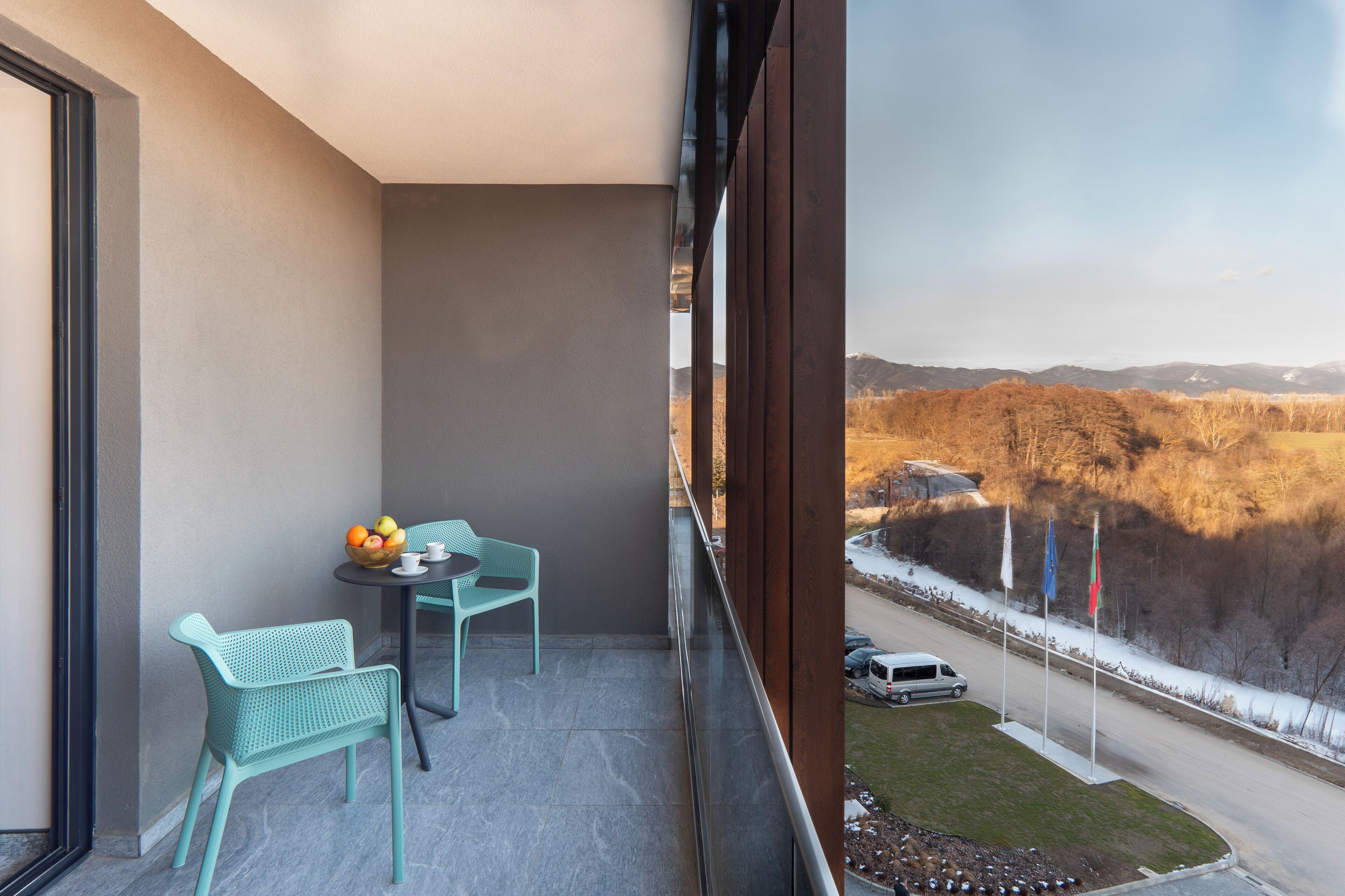 A balcony with two chairs and a table, a view of a road with flags, and a mountainous landscape.