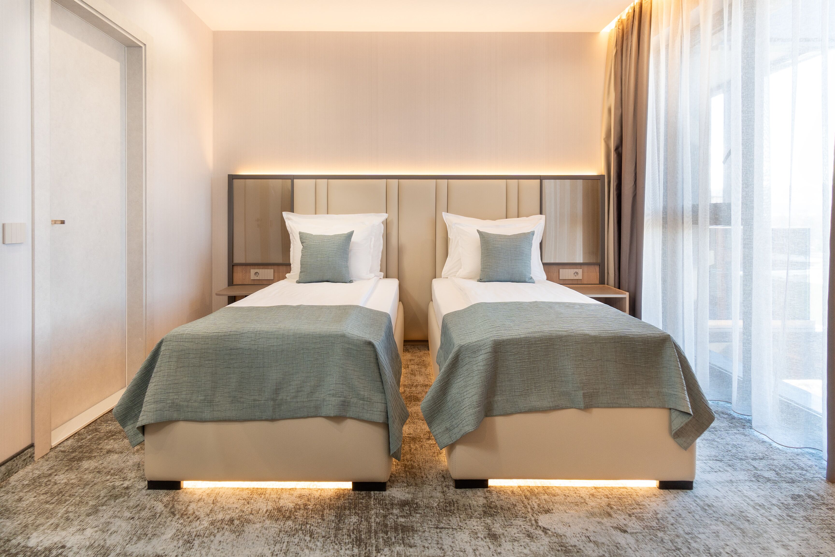 A hotel room with two beds, a headboard with integrated lighting, a large window with sheer curtains, and a carpeted floor.