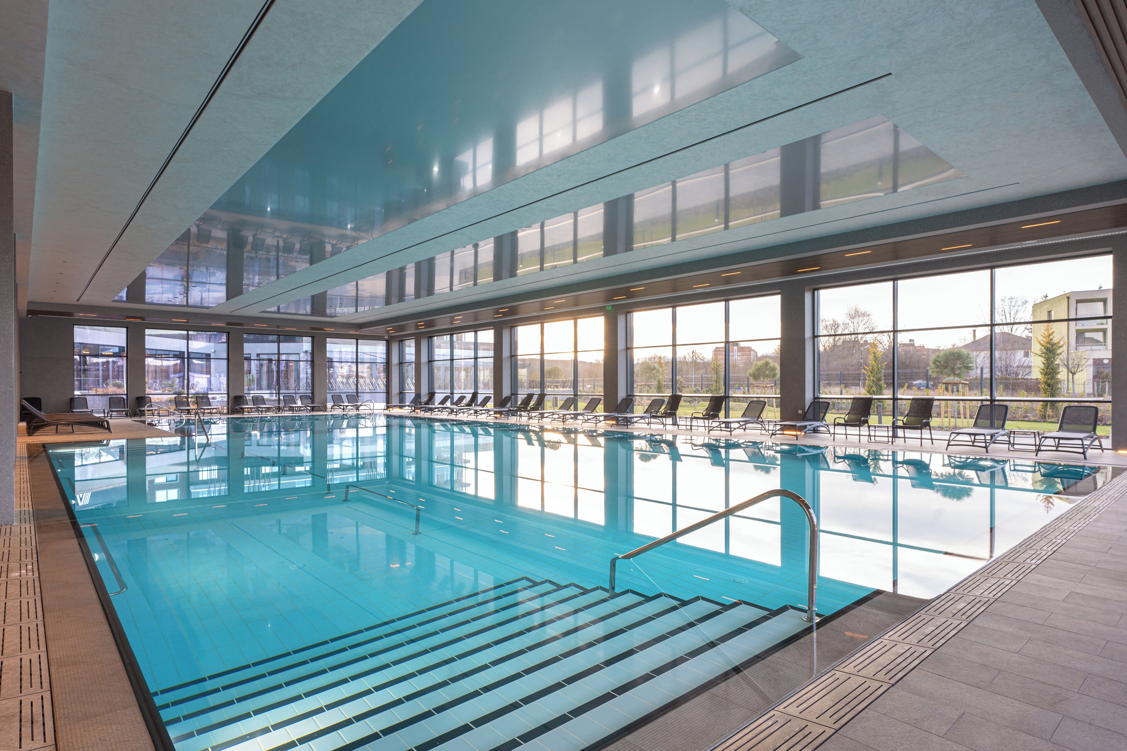 An indoor swimming pool with clear glass walls, multiple lounge chairs, and a view of the surrounding buildings.