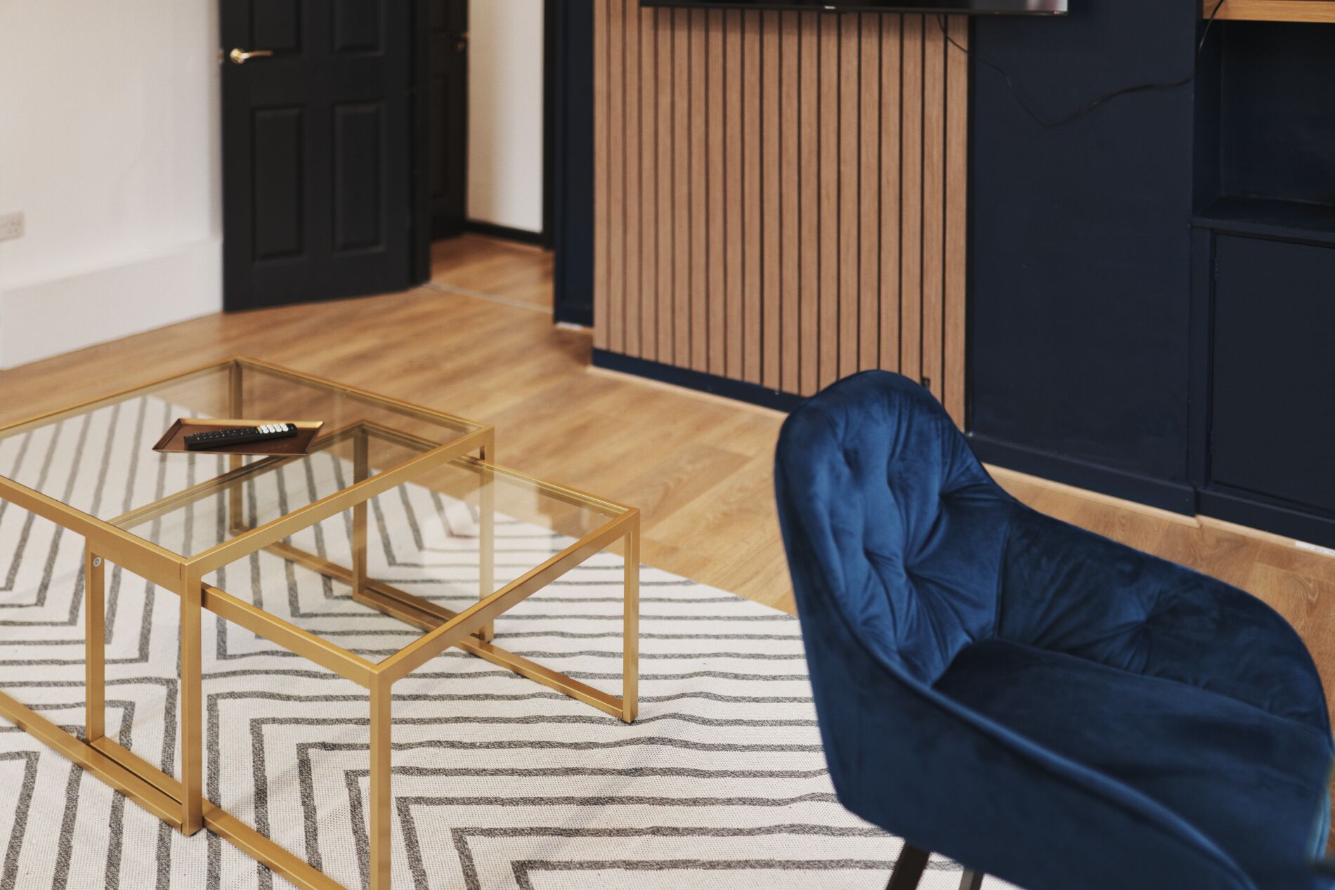 Una sala de estar con una mesa de centro de vidrio, un sillón azul, un tapete a rayas y piso de madera.