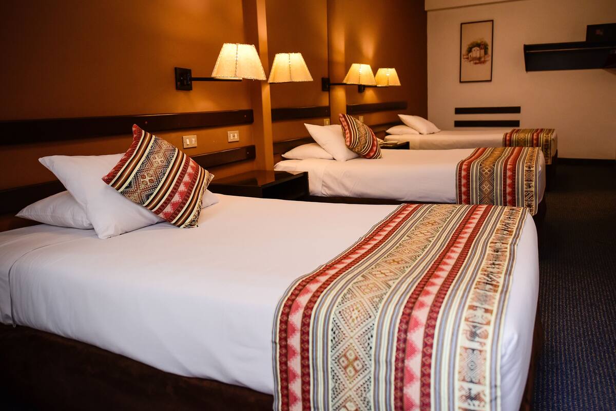 Quarto de hotel com três camas, cada uma com colcha e almofadas estampadas. Há luminárias fixadas na parede acima de cada cama.
