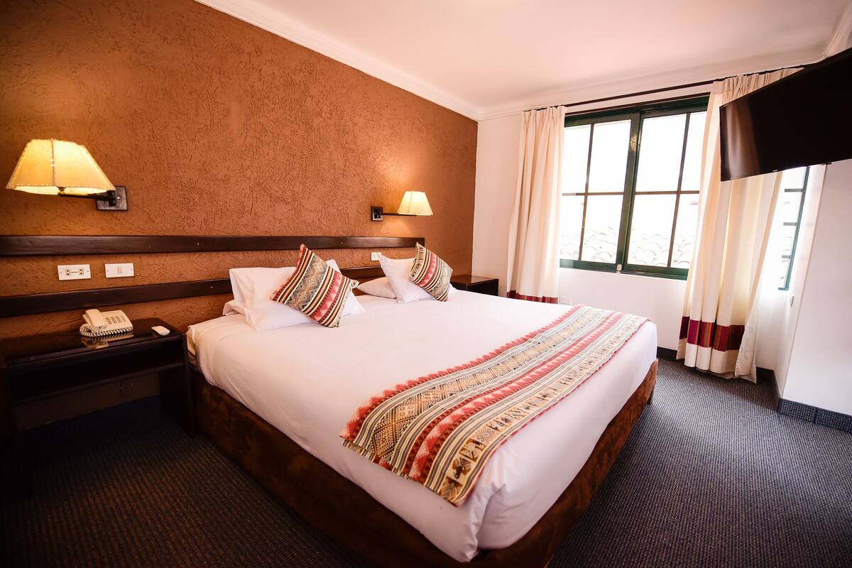 Quarto de hotel com uma cama grande, duas luminárias de cabeceira, um telefone e uma janela com cortinas.