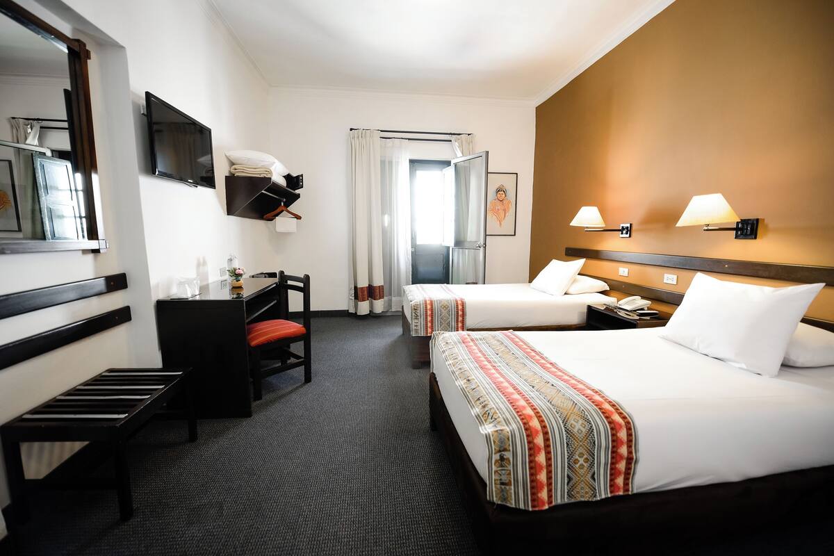 Quarto de hotel com duas camas, uma mesa de trabalho com cadeira, televisão e banheiro com cortina de chuveiro.