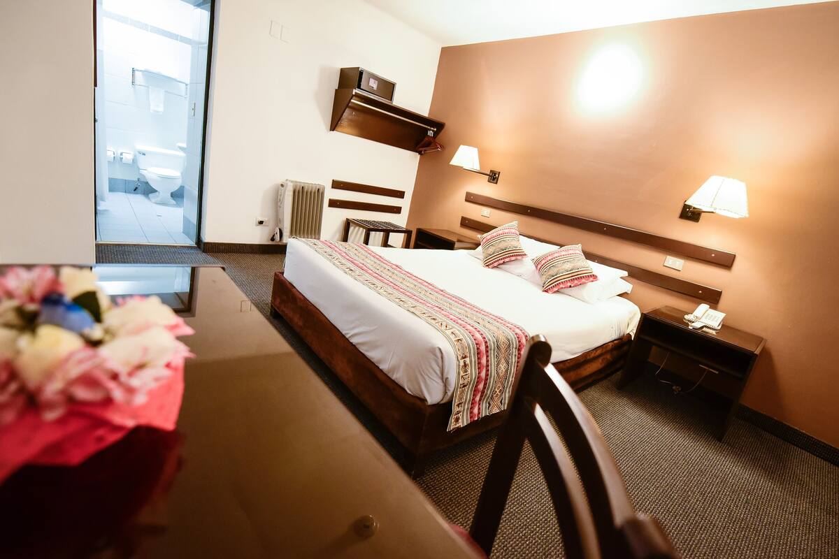 Quarto de hotel com cama, mesas de cabeceira, escrivaninha e uma cadeira.