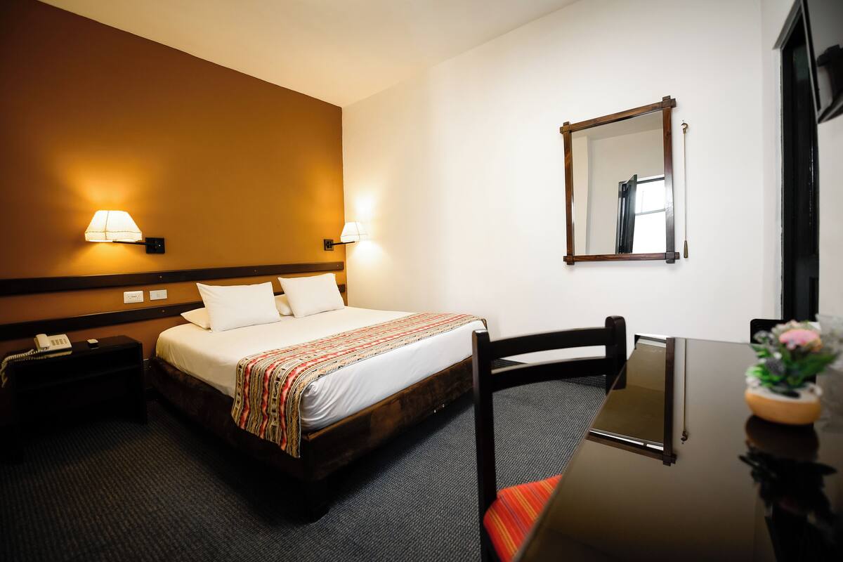 Quarto de hotel com uma cama, abajures, uma escrivaninha com cadeira e um espelho na parede.