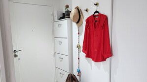 A hallway with a white door, a grey bench with a brown bag, a red shirt on a hanger, and a white wardrobe with drawers. - Sublime Appartement Proche de Paris et La Défense Arena (Courbevoie)