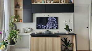 A modern living room with a flat-screen TV mounted on the wall, wooden cabinets, a black marble countertop, and various decorative items including plants and a zebra painting. - Sublime Appartement Proche de Paris et La Défense Arena (Courbevoie)