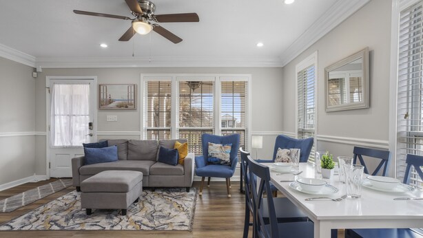 A dining area with a white table set for four, blue chairs, and a gray sectional sofa with blue and yellow pillows. - The Wave @ Coastal Retreat Villas (Long Beach)
