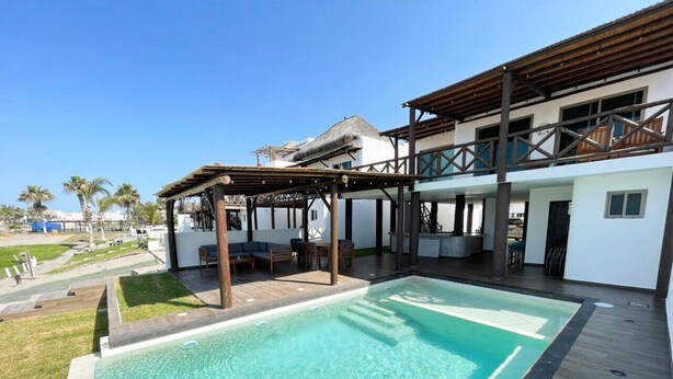Una piscina con cubierta de madera y zona de descanso, un espacio de estar cubierto, y un edificio con múltiples ventanas y balcones.