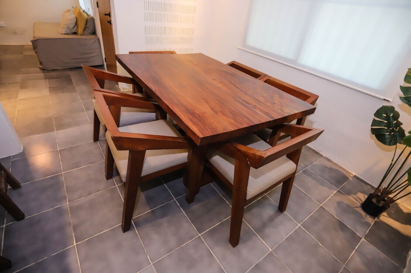 Un espace repas comprenant une table et des chaises en bois, un sol carrelé, un canapé et une grande plante.