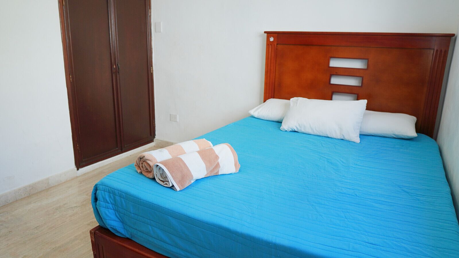 A bed with a blue bedspread and white pillows, a wooden headboard with cut-out panels, and a rolled towel on the bed.