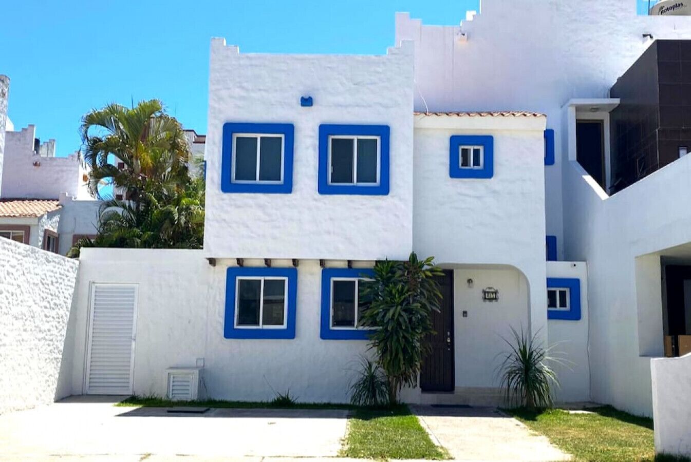 A white building with blue window frames and a door, surrounded by a small lawn and palm trees.
