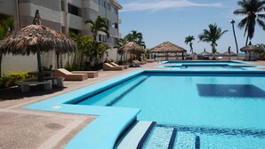 Una piscina con agua azul, rodeada de palmeras y sombrillas de paja.