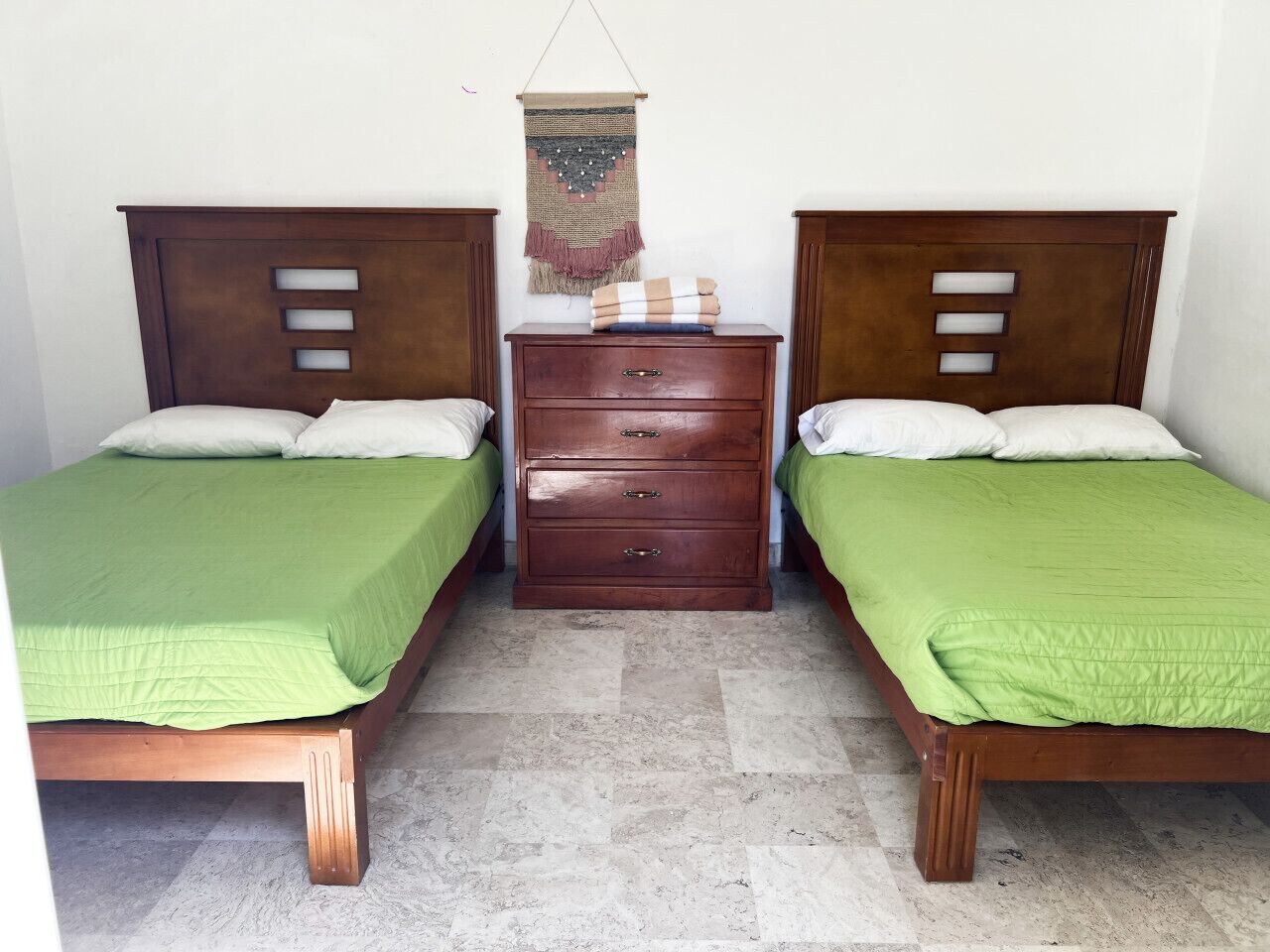 A room with two wooden beds, each with a green bedspread and white pillows, a wooden nightstand with drawers, and a wall hanging above the nightstand.