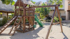 A wooden play structure with a slide and swings, set on sandy ground with palm trees and buildings in the background. - Casa en nuevo Altata, Punta Esmeralda Grand (Nuevo Altata)
