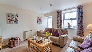 Ein gemütliches Wohnzimmer mit einem beigefarbenen Sofa, einem Holztisch und einem Fenster mit floralen Vorhängen.