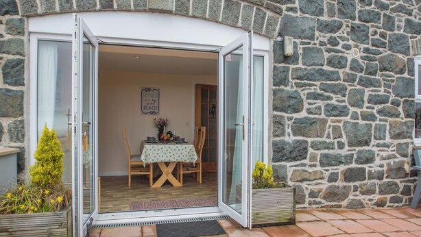 Ein Steinhaus mit Glastür und Eingangsbereich, der in einen Essbereich mit Tisch für zwei Personen, einem kleinen Topfpflanzen und einem gerahmten Bild führt.