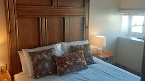 Un lit bien fait, avec du linge de lit blanc et des coussins décoratifs, une tête de lit en bois, une lampe de chevet et une fenêtre laissant entrer la lumière du jour.