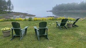 Blick auf einen See mit drei Adirondack-Stühlen, einer Wiese, gelben Blumen und ruhigem Wasser.