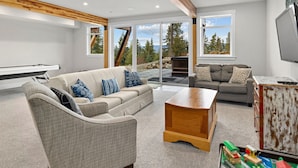 A modern living room with a sofa, a coffee table, and a television. There is a sliding glass door leading to an outdoor area with a view of trees and a mountain. - Huge game room, Hot tub, Sleeps 16+ with Bonus loft, ATV Snowmobile Trails (Cle Elum)
