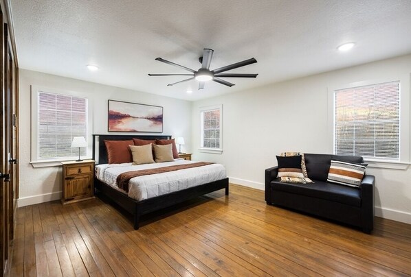 A bedroom with a bed, a black sofa, wooden flooring, ceiling fan, and two windows with blinds. - Creekside Ranch - Big Yard, Hot Tub, Fire Pit, Game Room, Pet-Friendly (Broken Bow)
