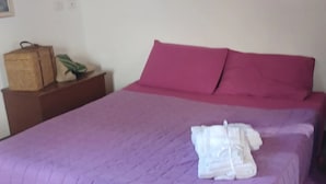 Ein Zimmer mit einem großen Bett, violetter Bettwäsche, einem Wanddekor und einem Nachttisch aus Holz mit Korb und Pflanze.