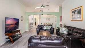 A living room with a flat-screen TV, leather sofas, a coffee table, and a ceiling fan. - Cozy Oasis 5BR Near Disney with private pool (Kissimmee)