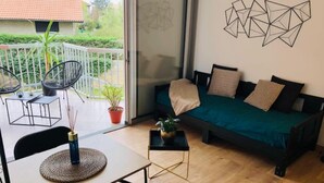 A cozy living area with a sofa, a coffee table, and a dining table. There is a balcony with chairs and a potted plant.