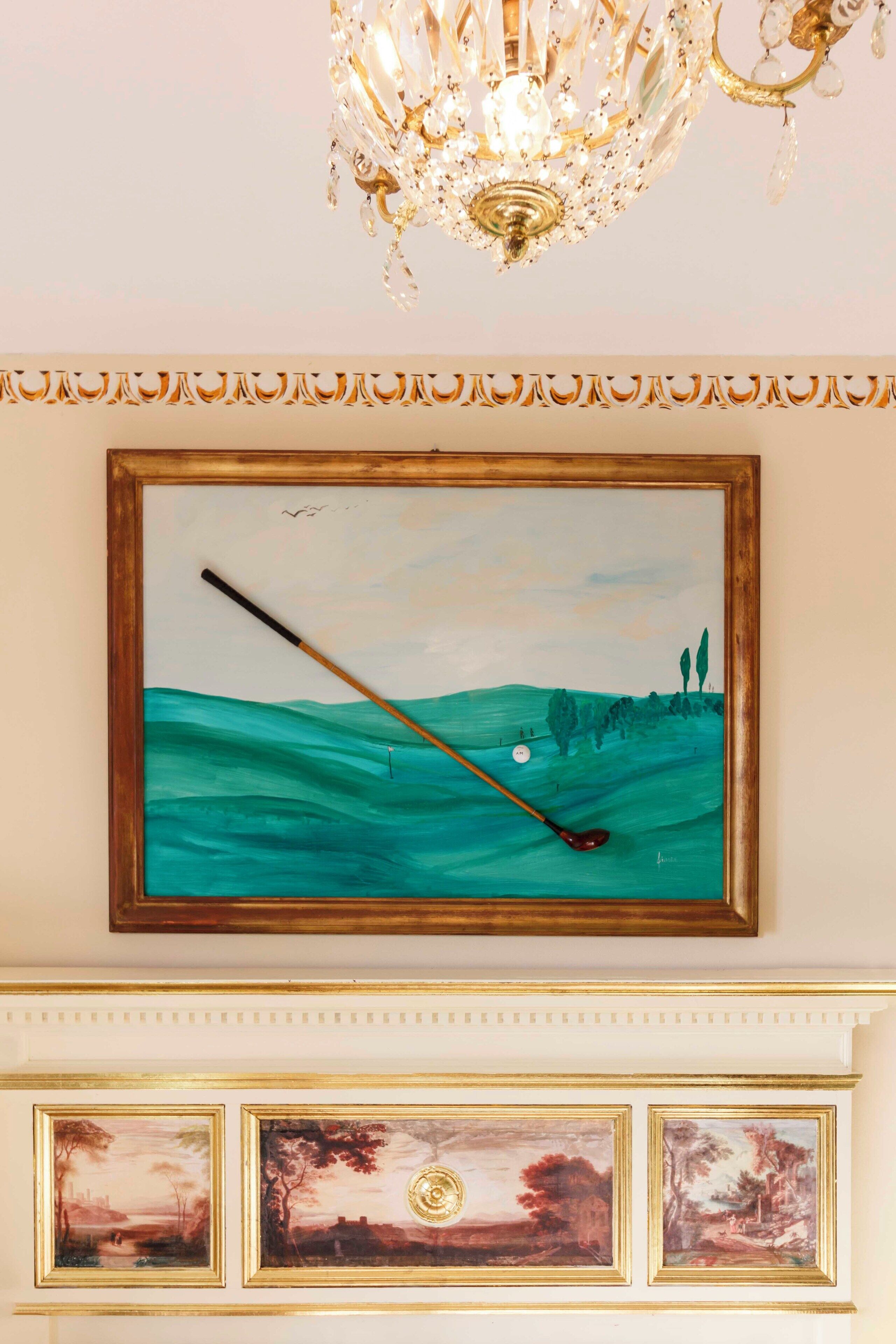 Un tableau encadré représentant un terrain de golf, avec un club de golf et une balle, placé au-dessus d’une cheminée ornée de trois tableaux plus petits.