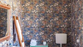 Un baño con papel tapiz floral, un lavamanos blanco, un inodoro, un estante verde y una araña.