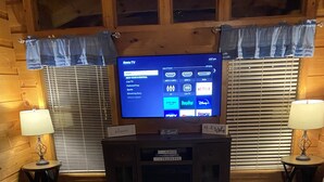 A cozy living room with a large flat-screen TV mounted on a wooden wall, a small round table with a placemat, and two side tables with lamps. - Cozy quiet romantic cabin for 2, Fully equipped modern kitchen. Pleasant patio. (Sevierville)